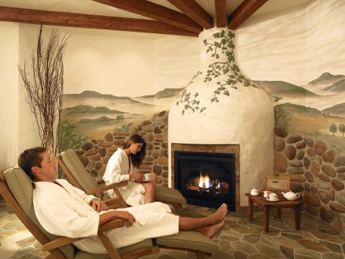 Two people in bathrobes relax in a cozy spa room with a fireplace and scenic wall murals.
