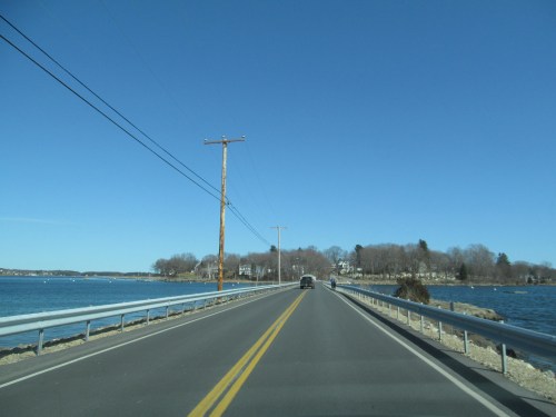A clear road stretches alongside a body of water, with power lines and trees visible under a bright blue sky.
