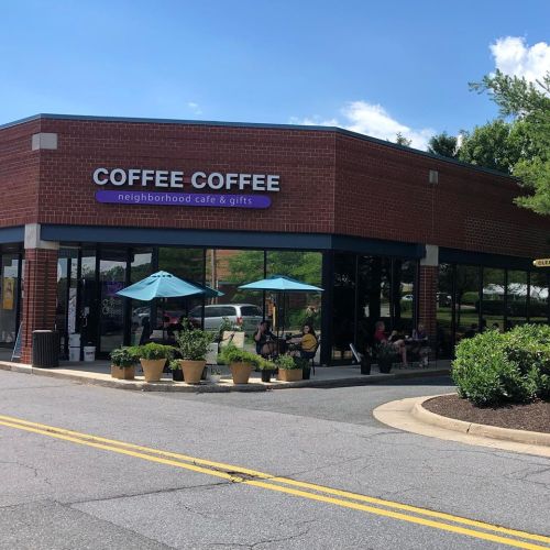 A brick café with outdoor seating and umbrellas, surrounded by greenery, on a sunny day.