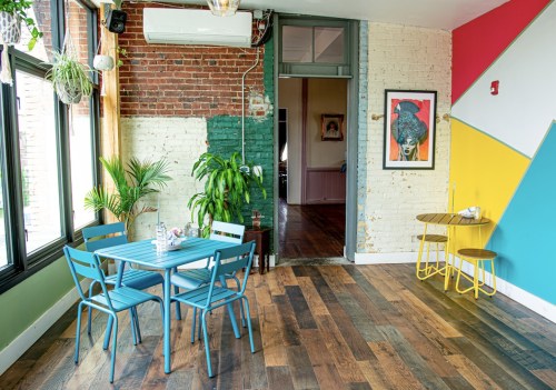 A bright, modern cafe interior with a blue table and chairs, plants, and colorful wall accents.