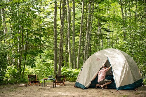 A person enters a camping tent in a lush green forest, with two chairs and a small table nearby.
