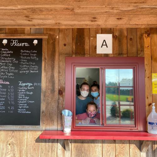 Three people wearing masks smile from a window of a rustic stand with a menu board listing flavors and prices.