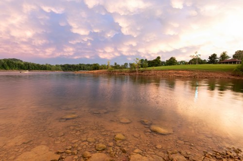 A serene river scene at sunset, with calm waters reflecting colorful clouds and a green shoreline in the background.