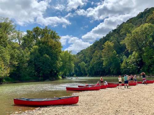 A serene river scene with red canoes on the shore, surrounded by lush green trees and a partly cloudy sky.