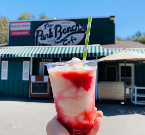 A hand holds a colorful frozen drink with a straw in front of The Park Bench Café, featuring a green and white striped awning.