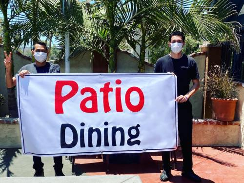Two masked individuals hold a large sign that reads "Patio Dining" in a sunny outdoor setting with palm trees.
