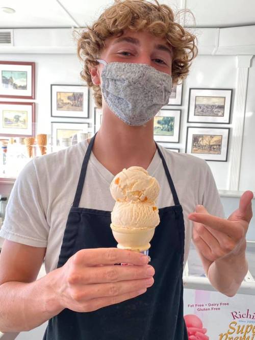 A young man in a mask holds a triple scoop ice cream cone, smiling and pointing with his other hand.