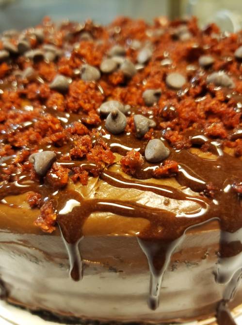 A close-up of a chocolate cake topped with chocolate chips and crumbled red frosting, drizzled with chocolate sauce.