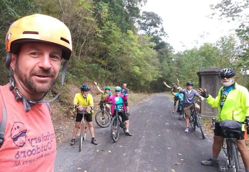 A group of cyclists poses on a forested road, smiling and waving, wearing helmets and colorful cycling gear.