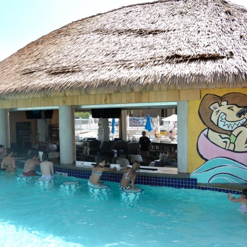 A swim-up bar with a thatched roof, people sitting on stools in the water, and a colorful mural in the background.