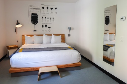 A modern hotel room featuring a bed with white linens, a mirror, and wall art depicting wine glasses.
