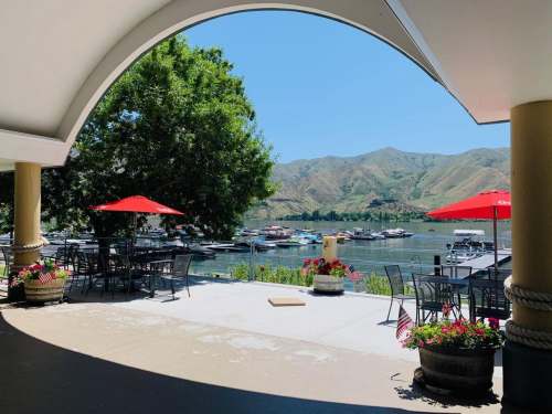 View of a lakeside patio with red umbrellas, boats on the water, and mountains in the background on a sunny day.