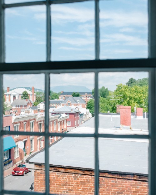 View through a window showcasing rooftops, trees, and distant hills under a clear blue sky.