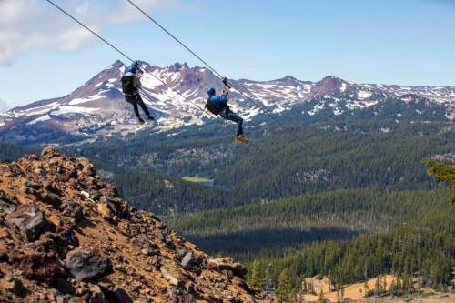 Two people ziplining over a mountainous landscape with snow-capped peaks and lush green forests below.