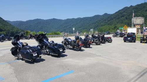 A row of motorcycles parked along a scenic highway with mountains in the background and flags lining the road.
