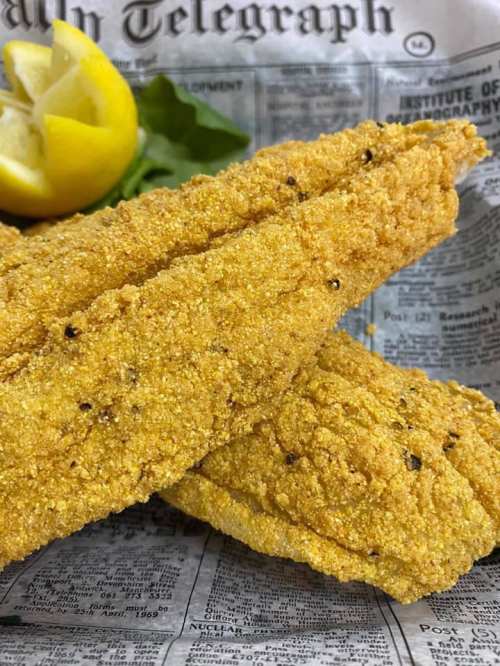 Two golden, crispy fried fish fillets on newspaper, garnished with a lemon wedge and fresh greens.