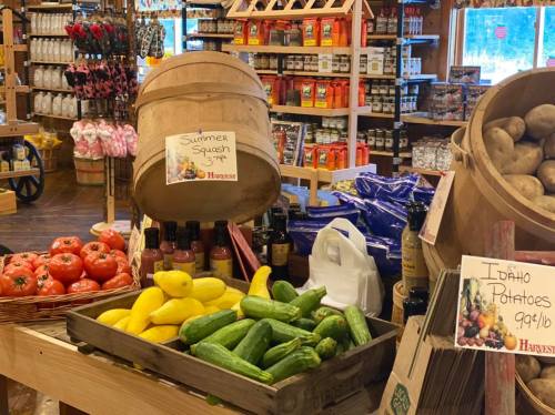 A rustic market display featuring fresh vegetables like summer squash, tomatoes, and Idaho potatoes, with various products in the background.