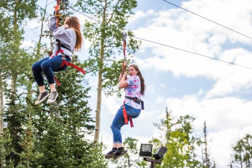 Two girls ziplining through a forested area, surrounded by trees and a blue sky with clouds.
