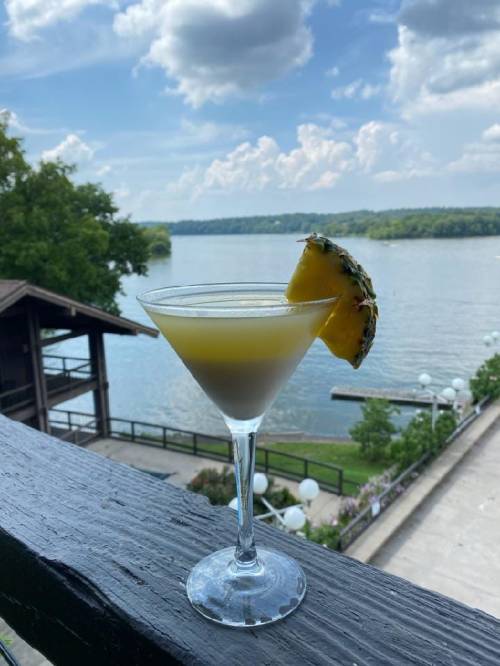 A tropical cocktail in a martini glass, garnished with a pineapple slice, overlooking a serene lake and blue sky.