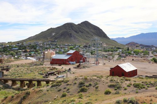 A mining town with red buildings and a mountain backdrop, surrounded by green hills and a clear blue sky.