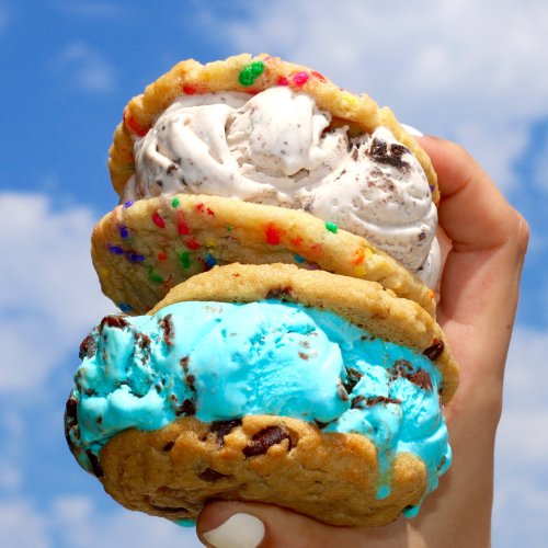 A hand holding two colorful ice cream sandwiches with cookie layers and vibrant ice cream flavors against a blue sky.