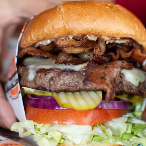 A close-up of a juicy burger with bacon, cheese, lettuce, tomato, pickles, and onions, held in a hand.