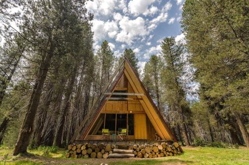 A modern A-frame cabin surrounded by tall trees under a partly cloudy sky. Logs are stacked in front.