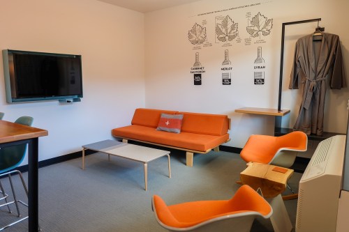 A modern hotel room featuring an orange sofa, two chairs, a TV, and wine-themed wall art.