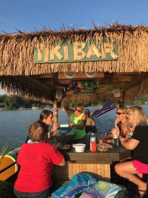 A group of people enjoying drinks at a tiki bar by the water, with a thatched roof and festive decorations.