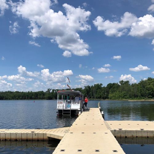 A boat is docked at a wooden pier on a calm lake, surrounded by green trees and a blue sky with fluffy clouds.