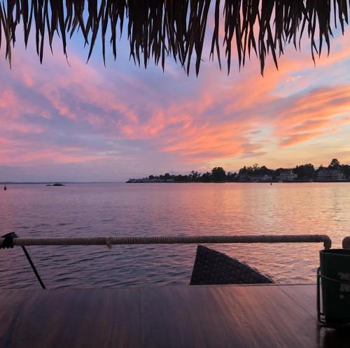 A serene waterfront view at sunset, with colorful clouds reflecting on calm water and a thatched roof in the foreground.