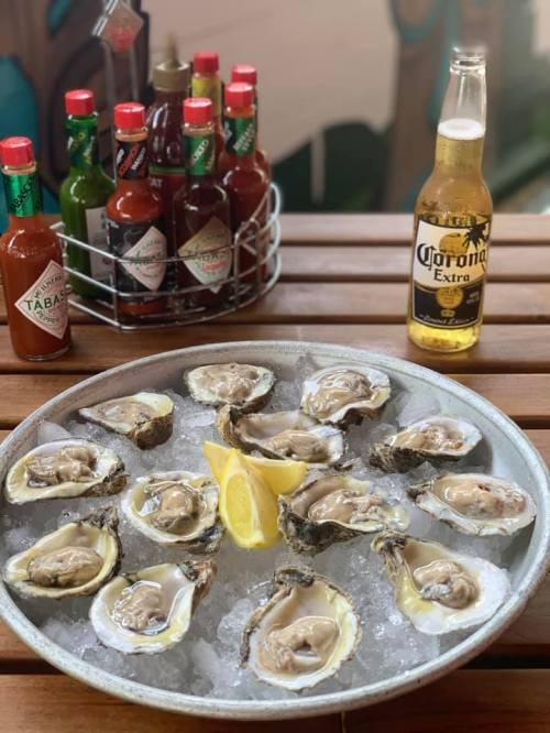 A platter of fresh oysters on ice, garnished with lemon, alongside a bottle of Corona and various hot sauces.