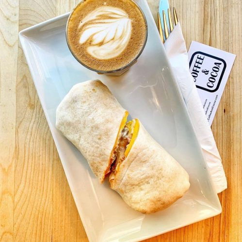 A plate with a wrapped burrito and a latte art coffee, set on a wooden table.