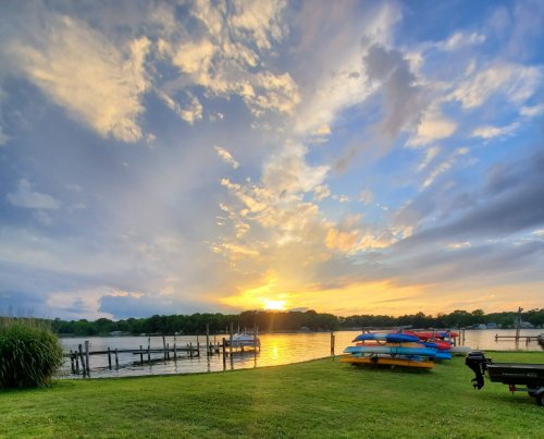 Sunset over a calm lake, with colorful kayaks lined up on the shore and clouds scattered in the sky.