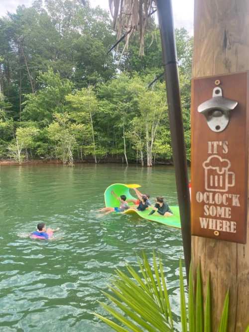 A group of people enjoying a sunny day on a colorful float in a lake, with a sign reading "It's O'Clock Somewhere."