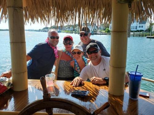 A group of five friends smiling together at a tiki bar by the water, wearing leis and enjoying a sunny day.