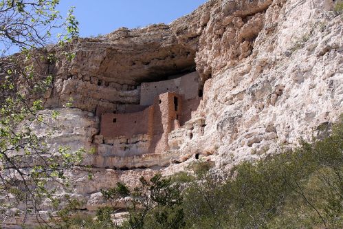 Ancient cliff dwelling nestled in a rocky overhang, surrounded by greenery and blue sky.