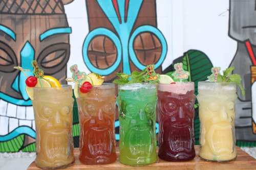 Four colorful tiki cocktails in decorative glasses, garnished with fruit and mint, set against a vibrant mural backdrop.