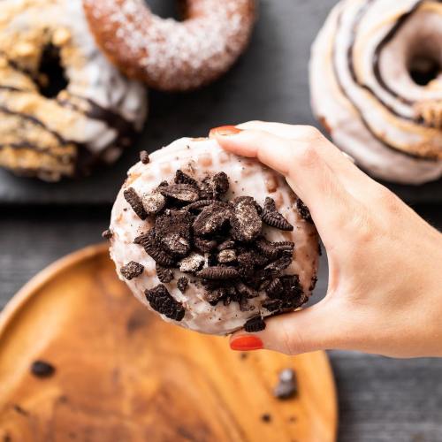 A hand holds a donut topped with crushed cookies, surrounded by other decorated donuts on a wooden plate.