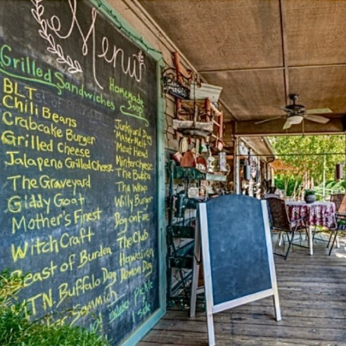 A rustic menu board displays various sandwich options outside a cozy eatery, with tables and decor in the background.