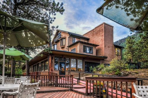 A wooden lodge surrounded by trees, featuring large windows and a deck with outdoor seating under green umbrellas.