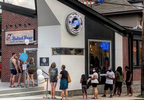 A line of people waiting outside a modern ice cream shop with a distinctive angled roof and colorful signage.