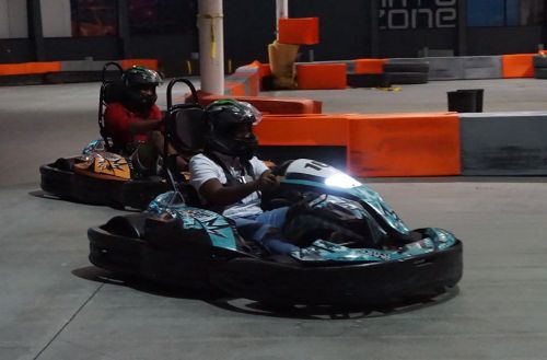 Two people racing go-karts on an indoor track, with colorful barriers and dim lighting in the background.