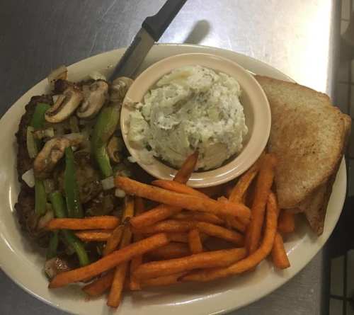 A plate with grilled steak topped with mushrooms and green peppers, sweet potato fries, potato salad, and toasted bread.