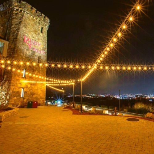 Night view of Ruby Falls with illuminated string lights and a stone tower, overlooking a cityscape.