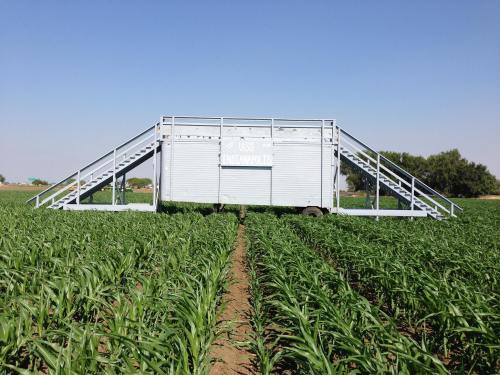 A large, mobile agricultural structure sits in a green field, surrounded by rows of crops under a clear blue sky.