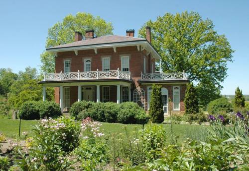 A historic brick house with a balcony, surrounded by lush greenery and colorful flowers under a clear blue sky.