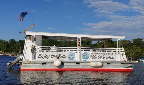 A white tour boat on calm water, featuring colorful flags and a sign for Slipaway River Tours with contact information.