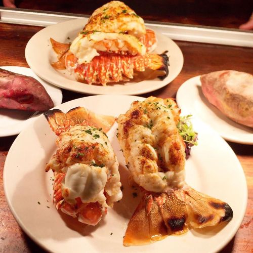 Two plates of grilled lobster tails, garnished with herbs, served alongside sweet potatoes.