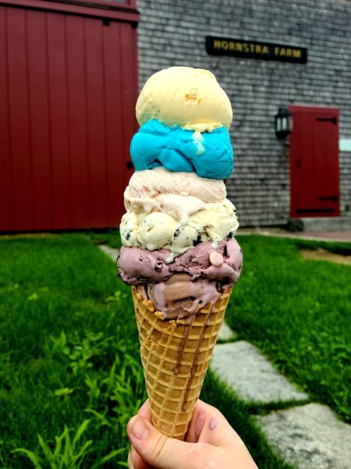 A hand holding a tall ice cream cone with four colorful scoops in front of a red barn.